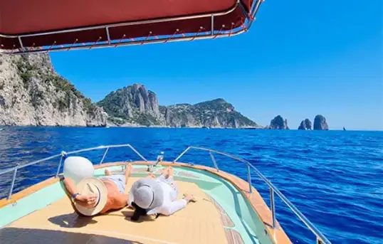A happy couple relaxing and sunbathing on the bow sundeck of the traditional Gozzo Sorrentino boat Bellavita, admiring the iconic Faraglioni rock formations during a private Capri boat tour with Frasil Charter