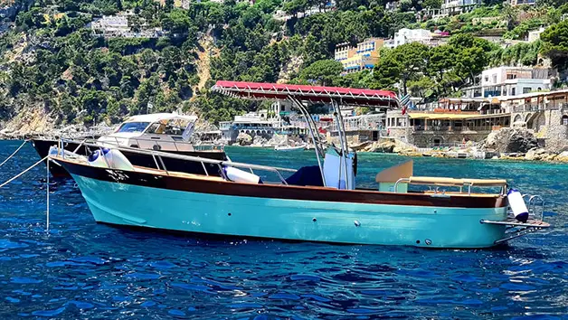 A high-angle drone view looking down at the spacious wooden deck of the Gozzo Sorrentino boat Bellavita anchored in the turquoise waters in front of Atrani.